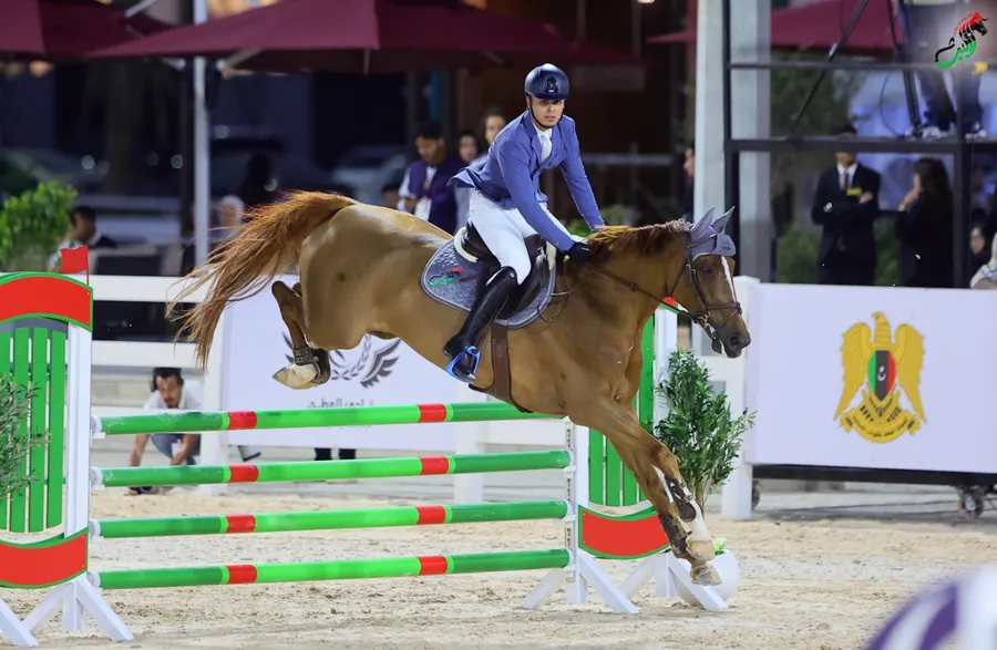 Chestnut horse clearing a green fence before the Libyan crest banner